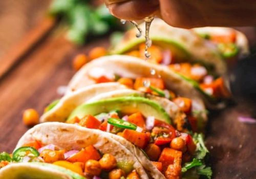 The image shows a hand squeezing lime over a row of fresh tacos filled with vegetables and chickpeas on a wooden board, garnished with lime slices.