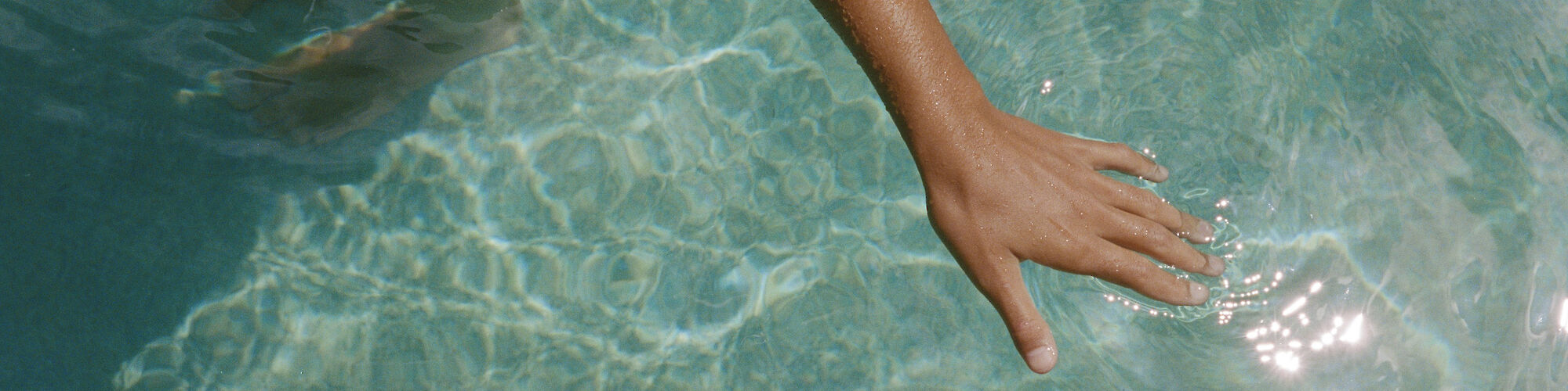 The image shows a person standing in a pool with clear water, one arm extended and hand touching the water; wearing a bracelet.