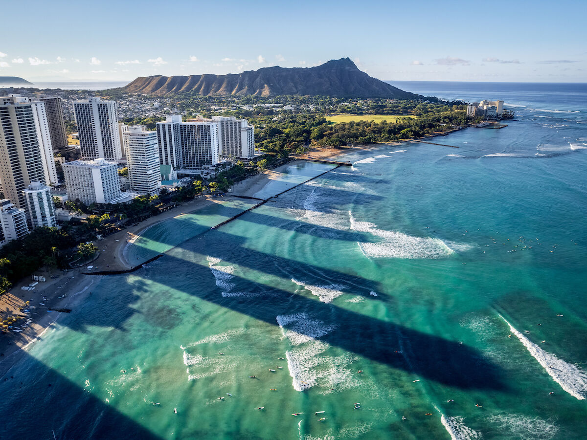 This image shows a coastal city with high-rise buildings along the beach, clear blue ocean, and a distinct mountain in the background, under clear skies.