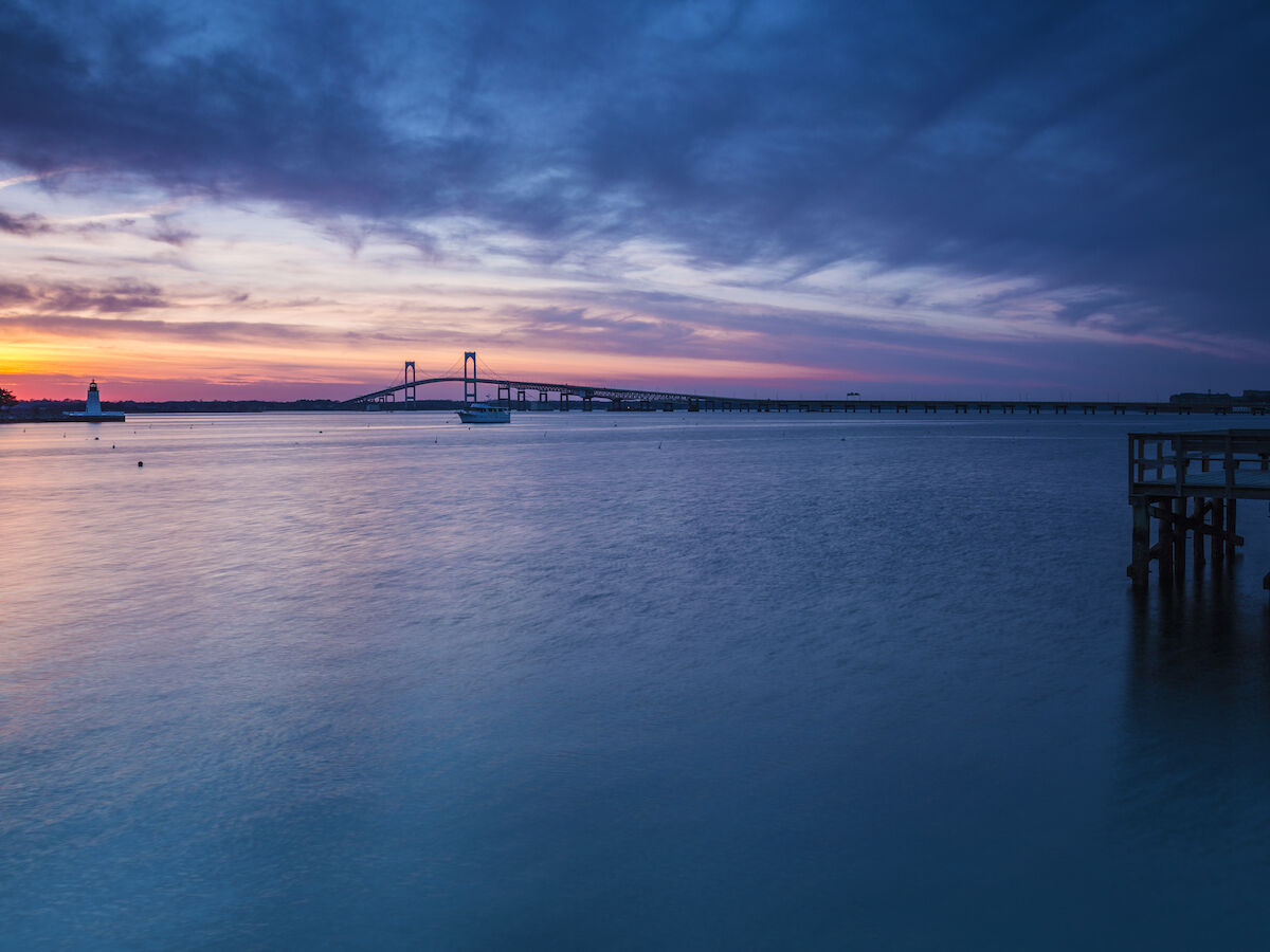 A tranquil sunset over a calm body of water, with a pier on the right and a distant bridge spanning the horizon under a dramatic sky.