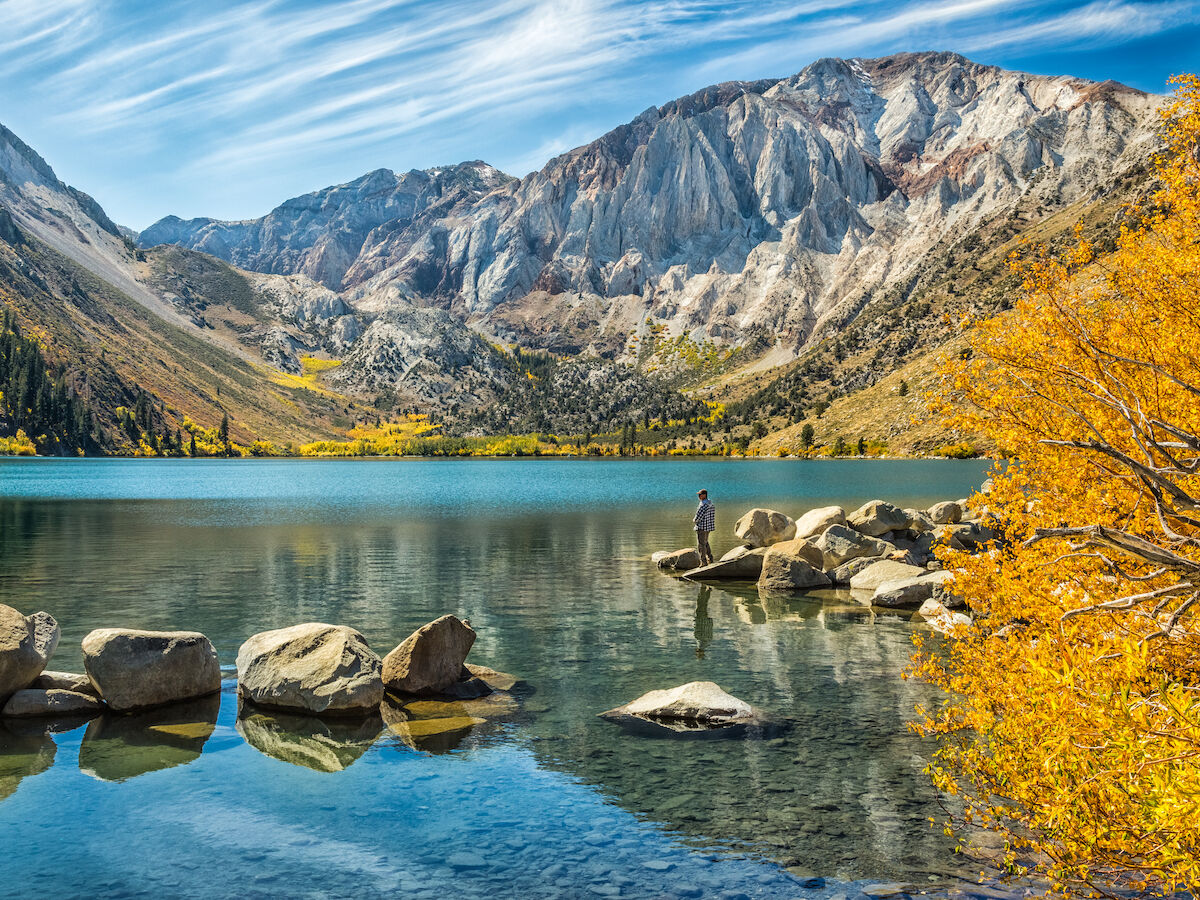 A serene lake is surrounded by rocky mountains and autumn trees. A person stands on rocks at the water's edge, reflecting on the calm surface.